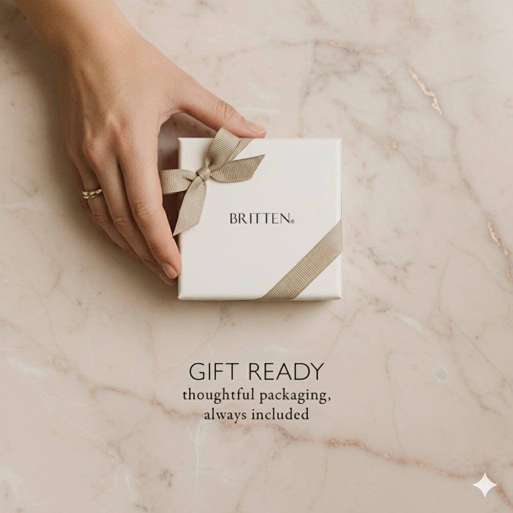 Hand holding a white gift box with a beige ribbon on a marble surface, featuring the brand 'Britten'.