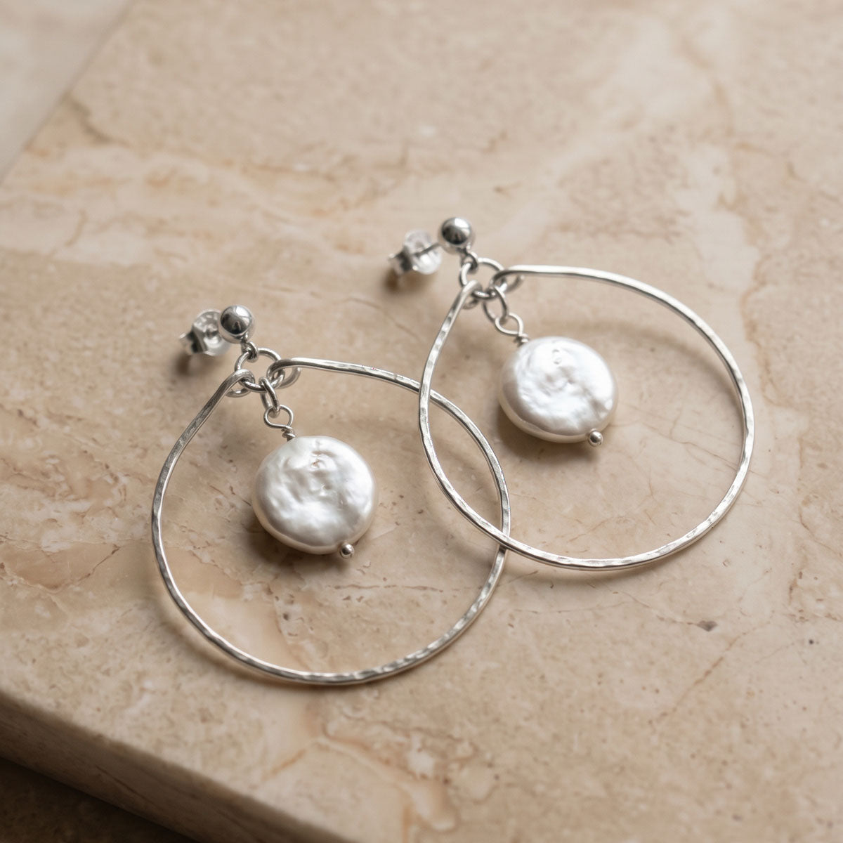 Sterling silver hammered teardrop hoop earrings with freshwater coin pearl drops, displayed on cream marble