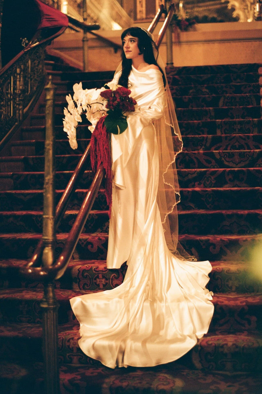 Dominique on a red staircase in a silk gown with a long train and a floor-length Juliet cap veil