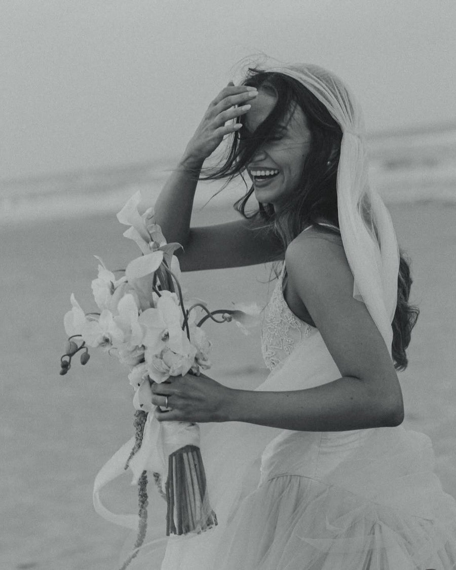 Rachel laughing on a windswept beach, hand in her hair, her Jules Juliet cap veil and orchid bouquet caught in the breeze