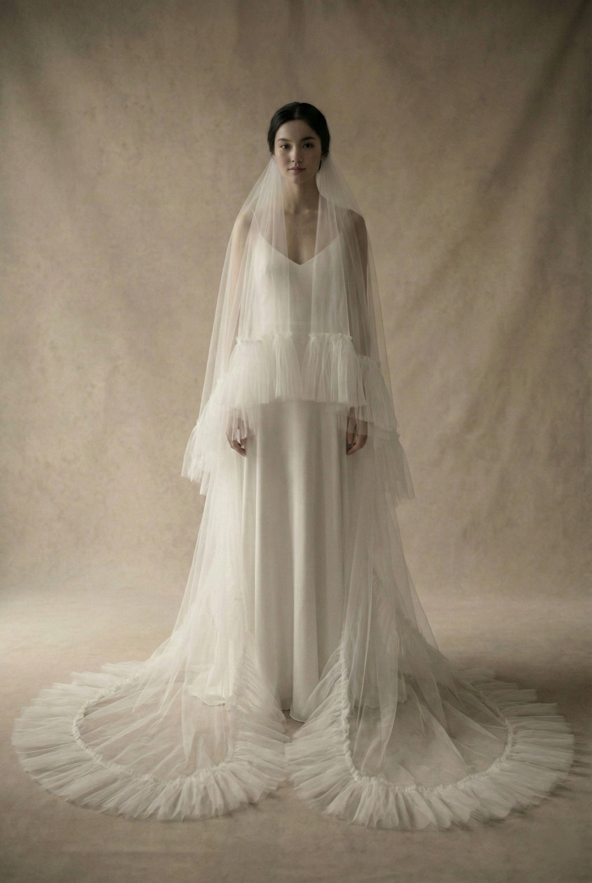 Full-length front view of a bride wearing a dramatic ruffle edge veil. The sheer blusher layer is worn over the face, while the long tier cascades down to pool on the floor, showcasing the textured, gathered tulle trim against a minimalist wedding dress.