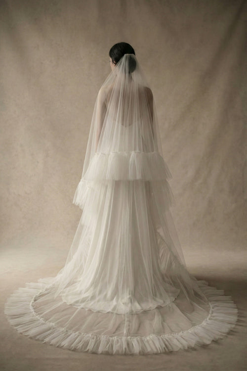 Back view of a voluminous two-tier wedding veil made from crisp traditional tulle. The design features a shorter blusher layer and a long cathedral train, both finished with a wide, gathered ruffle edge that creates a romantic, structural silhouette.